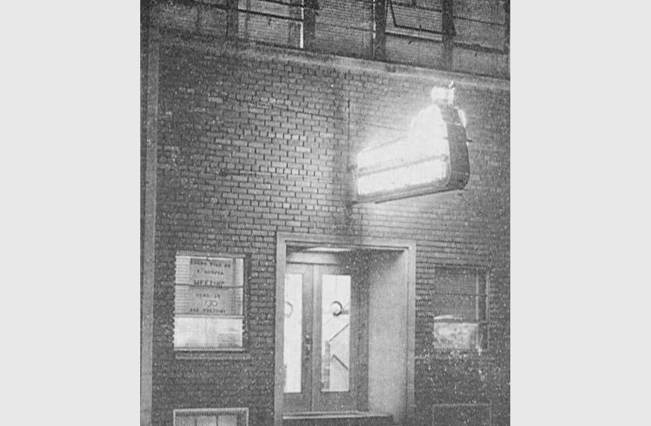 Historic brick storefront with a small lit sign projecting above the entrance and informational plaques mounted beside the doorway.