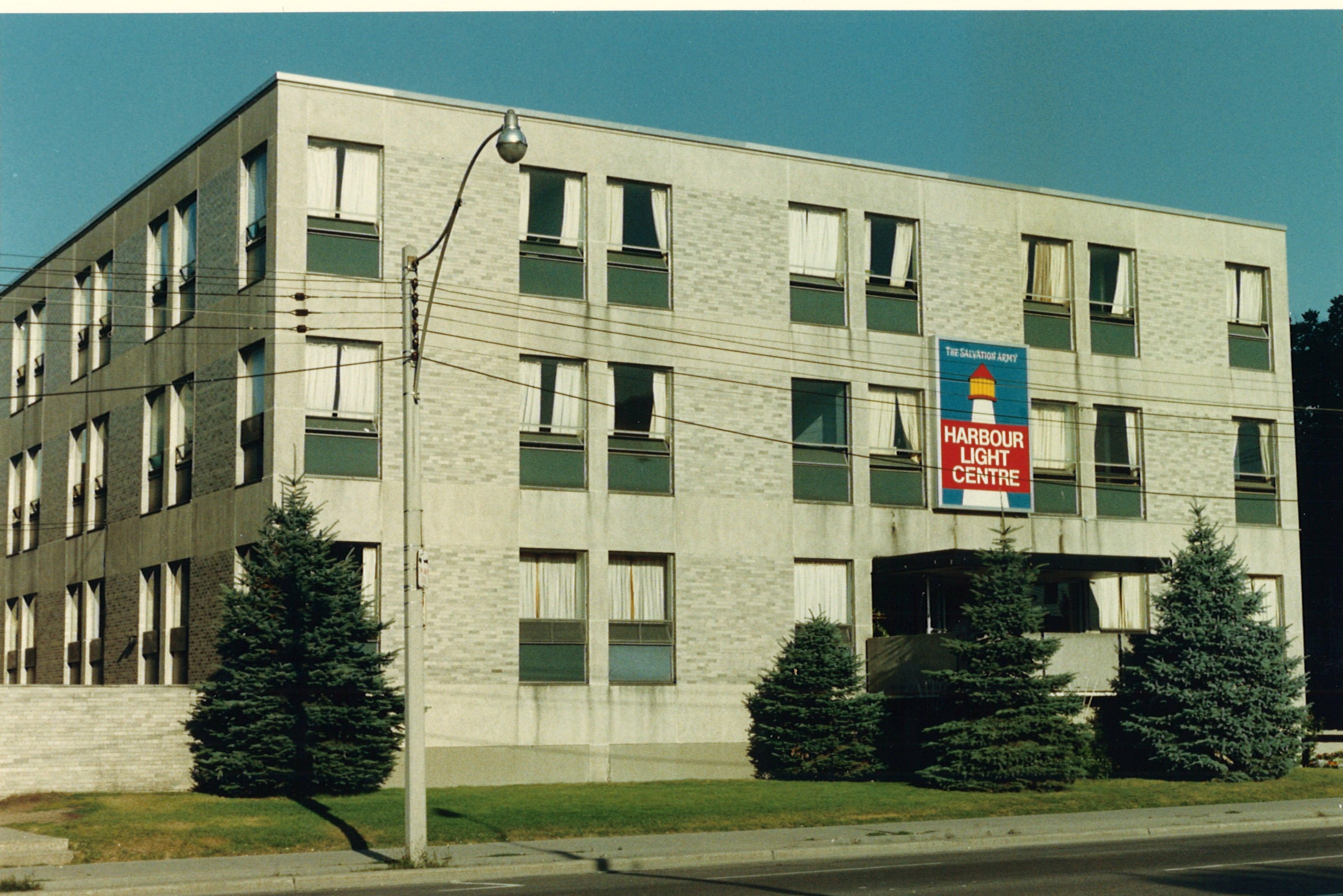 Exterior of the former Harbourlight building, a three‑storey structure with light brick walls, rows of square windows, evergreen trees along the front, and a Harbour Light Centre sign above the entrance.
