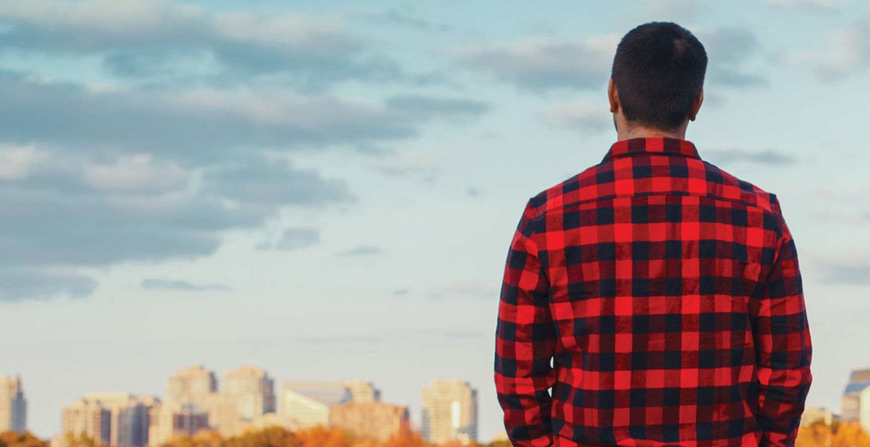 Individual wearing a red plaid shirt standing outdoors on a hilltop, looking over a city skyline under a cloudy sky.