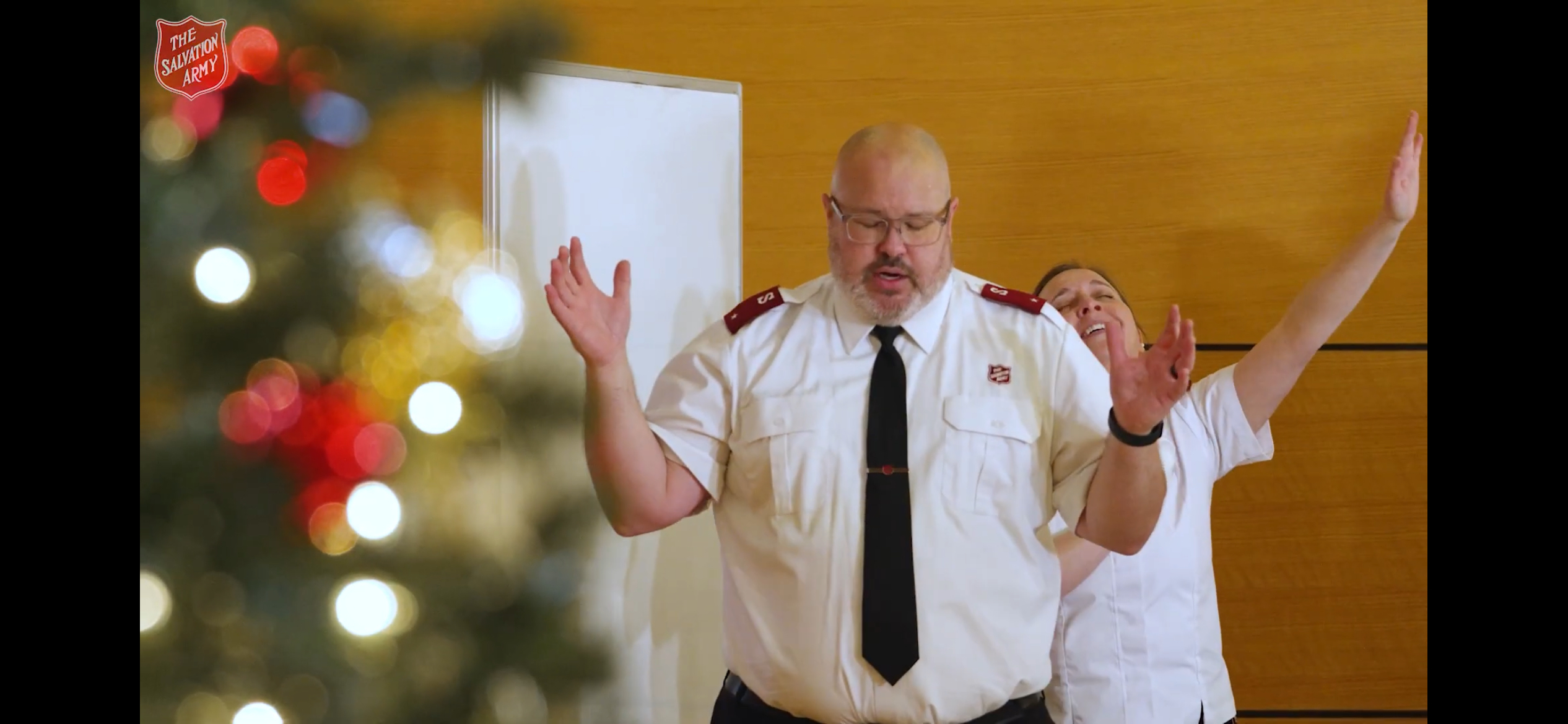 Salvation Army Corps Officers standing with raised hands in a worship setting at Downtown East Community Church, with a decorated Christmas tree in the foreground.