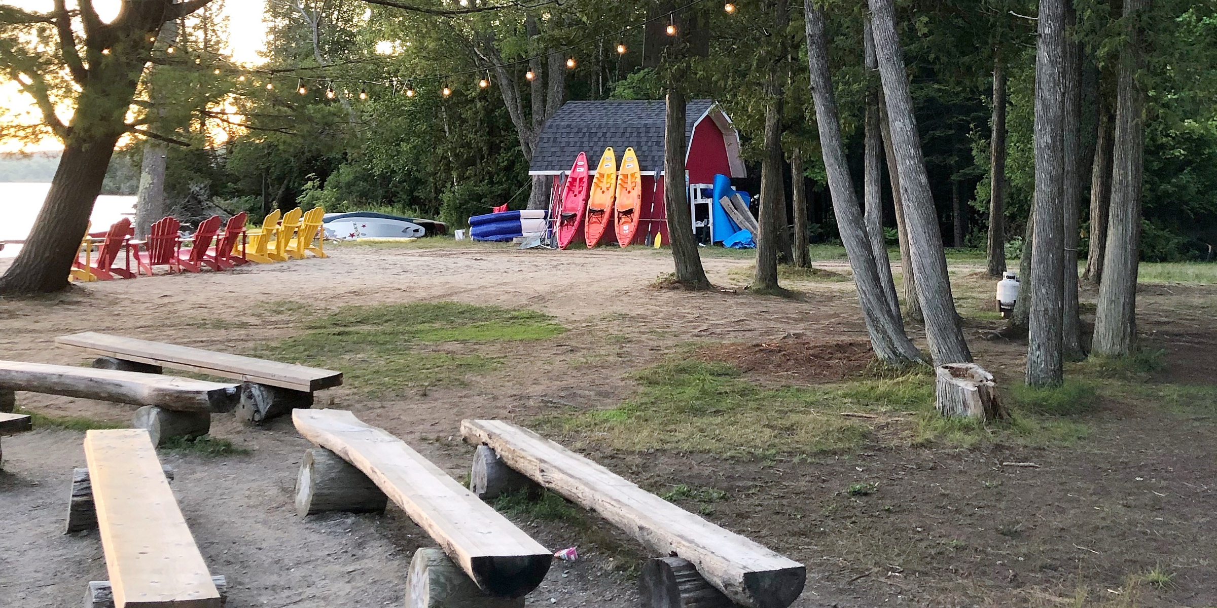 Outdoor camp area with wooden benches arranged around a fire pit, surrounded by trees and colourful chairs in the background.
