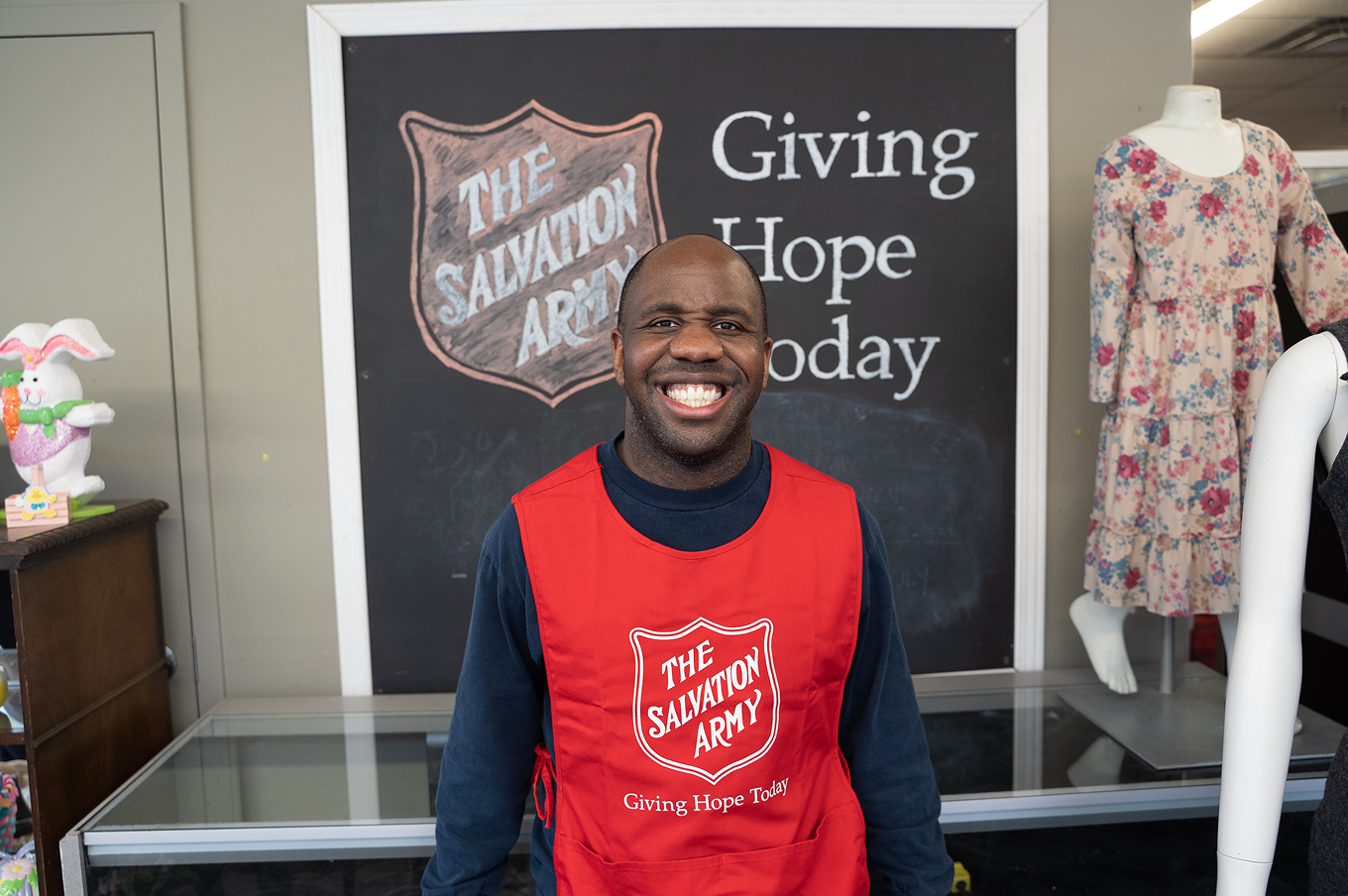 Smiling Salvation Army staff member wearing a red apron stands in front of a “Giving Hope Today” sign.